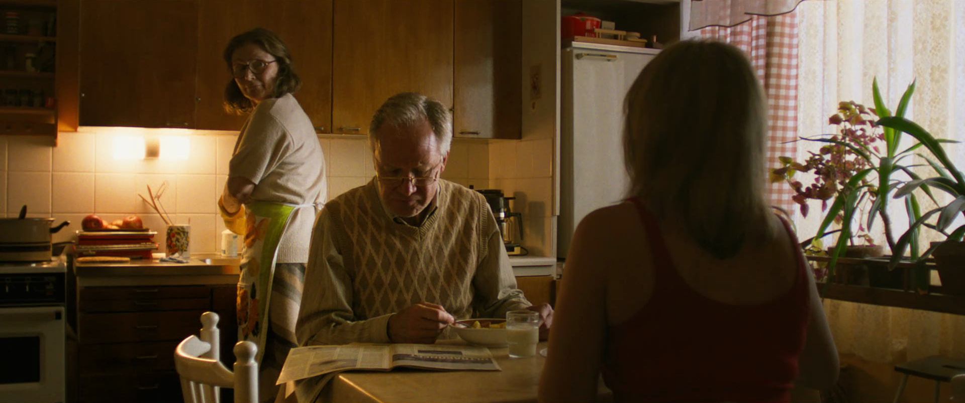 A family of four in a kitchen. A woman is in the background near the stove, a man is seated at the table eating, a woman with her back to the camera is sitting at the table, and a woman is sitting with her back to the camera on the right. The kitchen