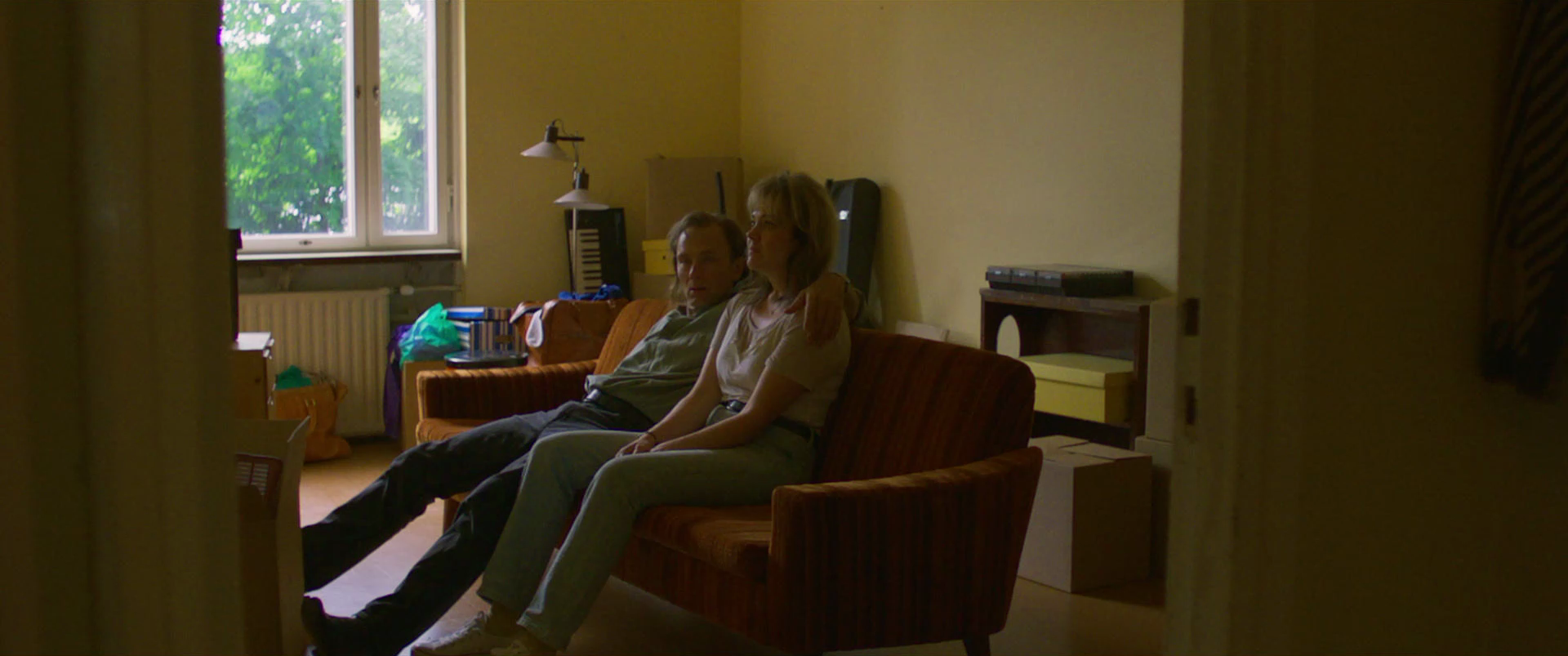 A man and woman sitting on a brown couch in a living room, with the man leaning back and the woman sitting upright, their arms around each other, near a window with green trees outside.