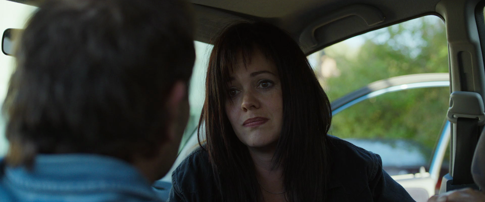 A woman with dark hair and bangs looking at a man with long hair inside a vehicle, with greenery visible through the window.