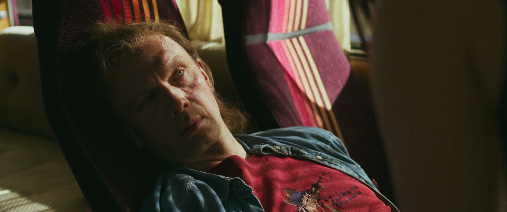 A man with long wavy hair reclining on a chair, looking upwards with a contemplative expression, surrounded by colorful striped fabric.