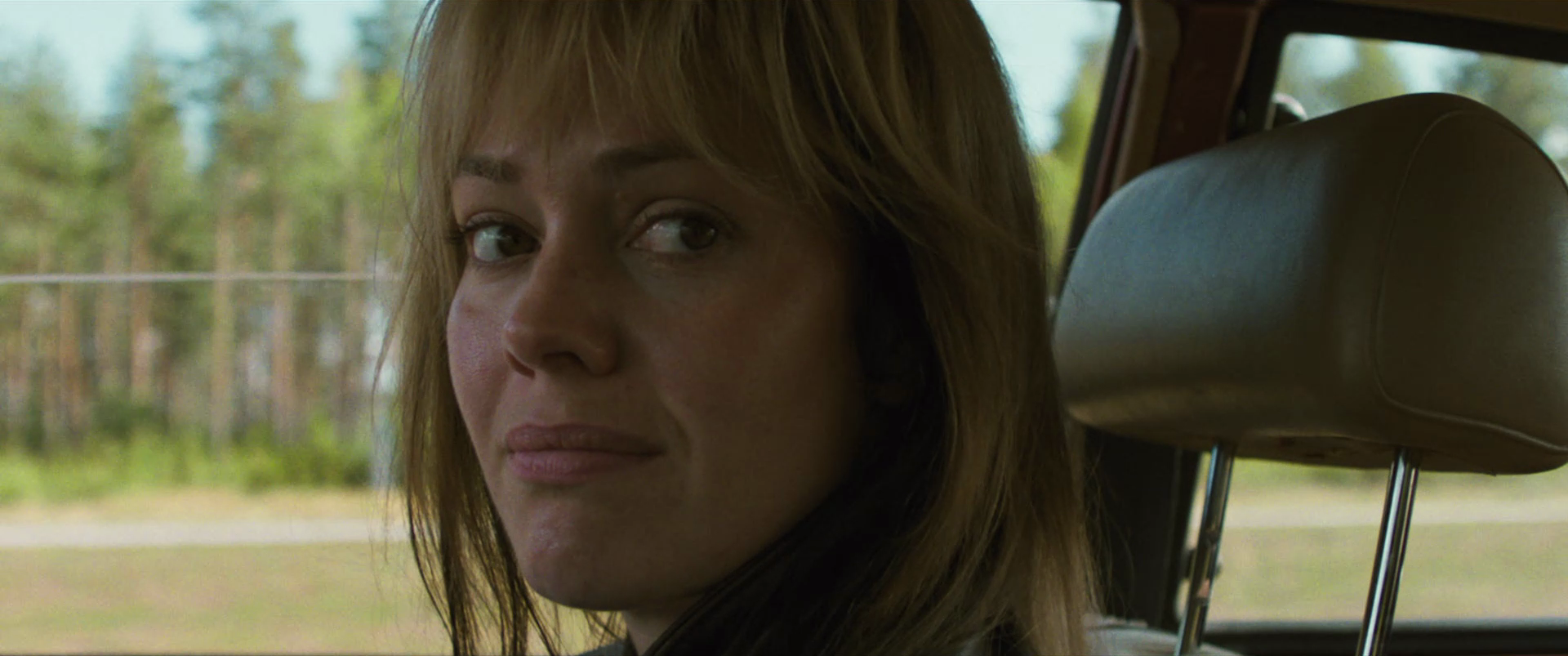 A woman with light brown hair sitting in the backseat of a car, looking out the window at a forested area with trees.