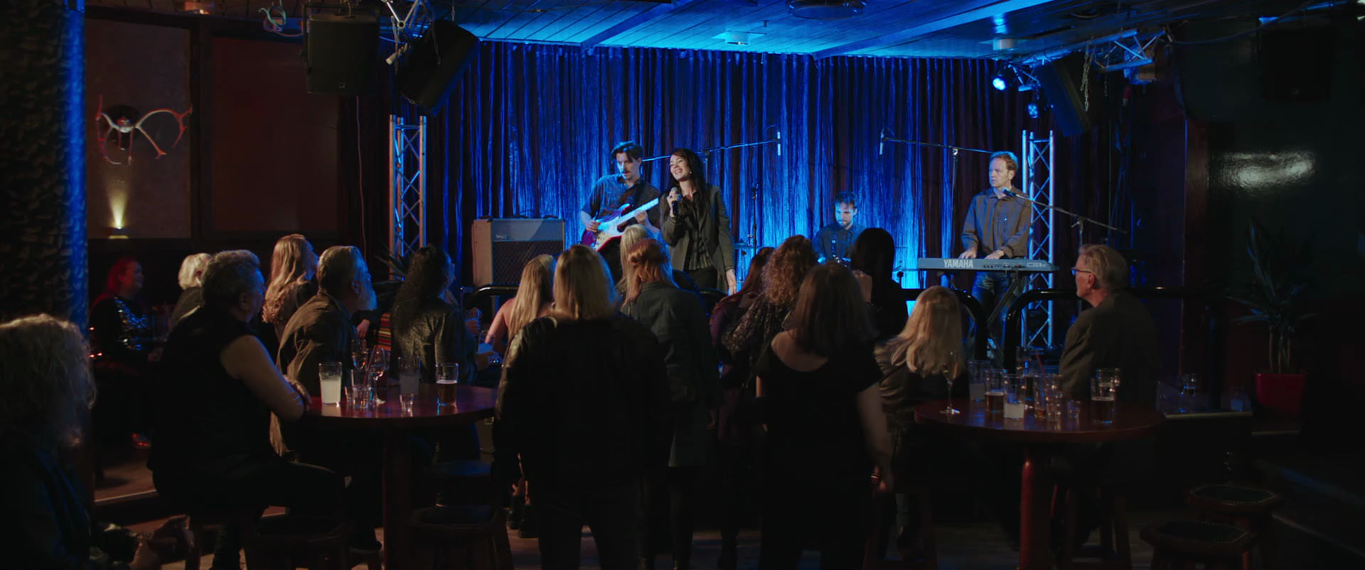 Live music performance on stage at a nightclub with a band and a singer, audience seated at tables, dim lighting, blue backlit curtain.