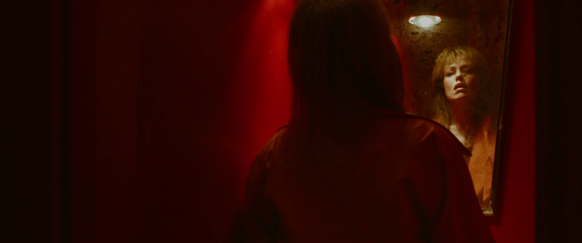 A woman with reddish hair looking at her reflection in a mirror, lit by a dim light, with red walls in the background.