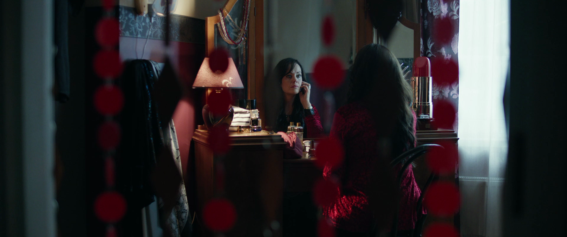 A woman sits at a vanity, looking into a mirror while talking on the phone. The scene is viewed through a doorway decorated with red beads, with a lamp, perfume bottles, and a mirror in the background.