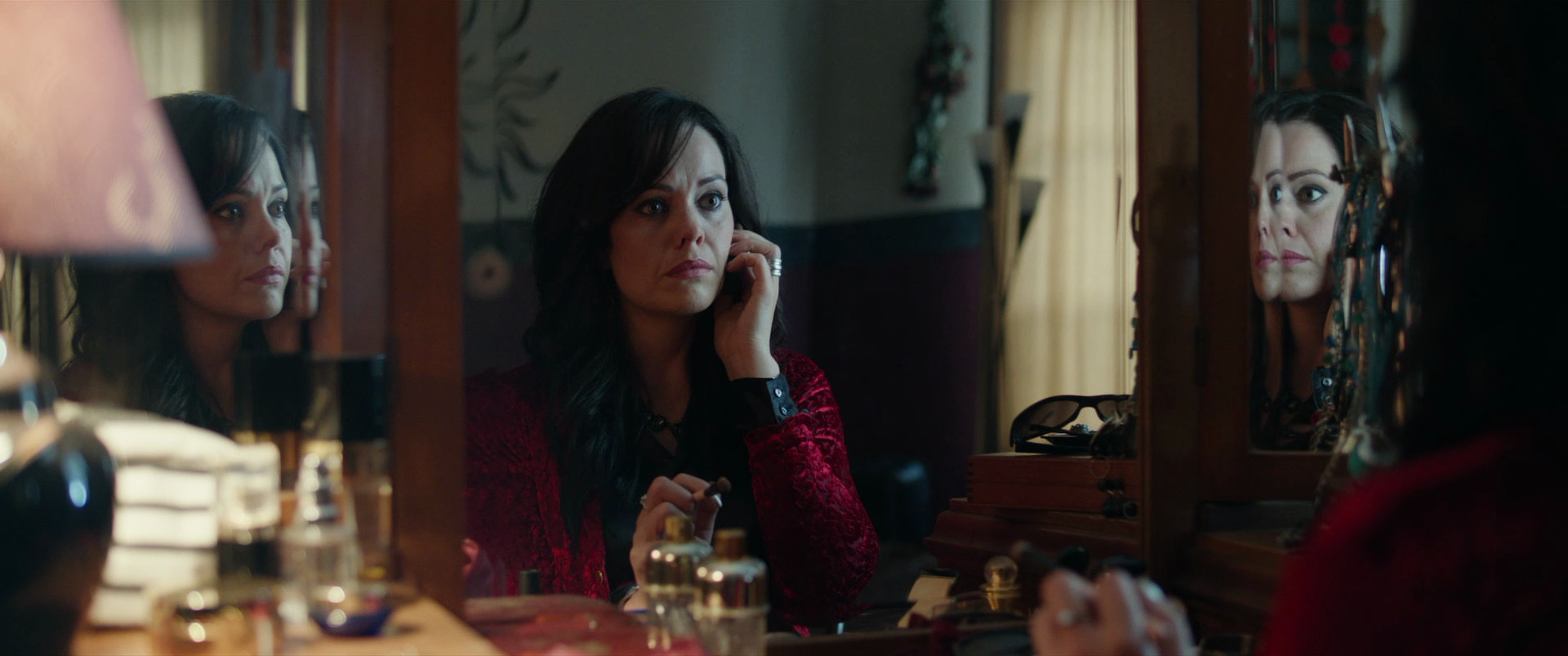 A woman with dark hair in a red jacket looks at her reflection in a mirror while putting on makeup, surrounded by beauty products and a jewelry box.