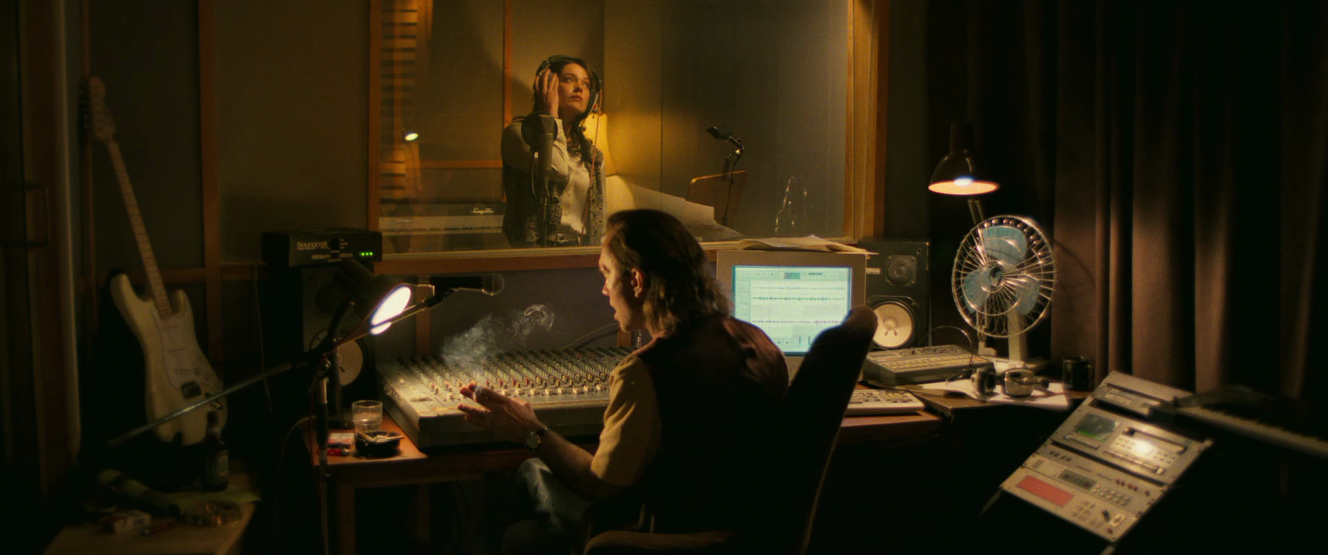 A woman in a recording studio wearing headphones on the other side of a glass window, and a man operating sound equipment in a control room with various audio devices, a guitar, and a fan.