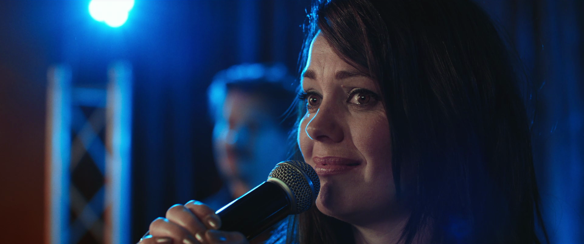 A woman with dark hair and makeup holding a microphone, appearing emotional, illuminated by blue stage lights, with a blurred background.
