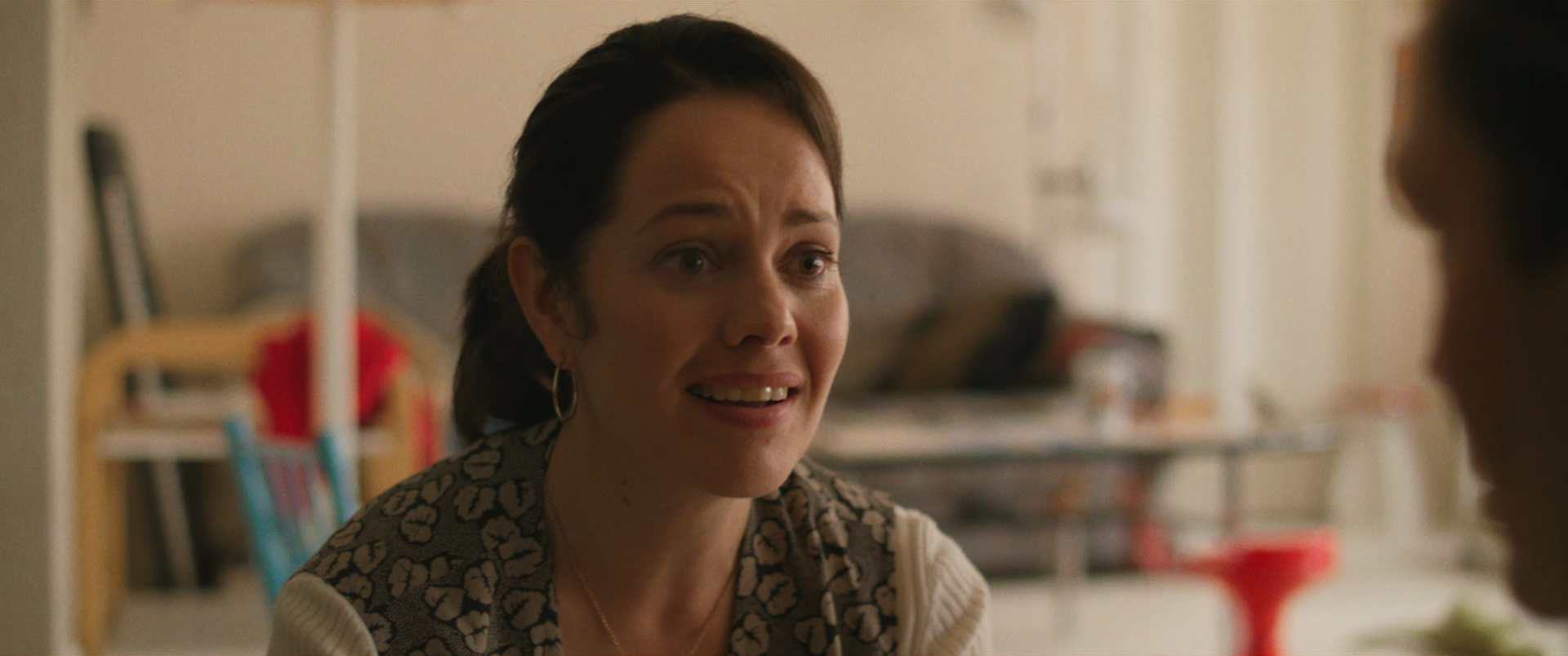 A woman with short dark hair, wearing a beige and brown patterned top and gold hoop earrings, appears to be talking or explaining something in a kitchen or living space.