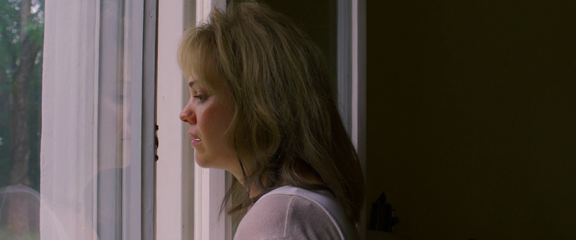 A woman with brown hair, wearing a light-colored top, looks pensively out of a window with white framing, while the room behind her is partially dark.