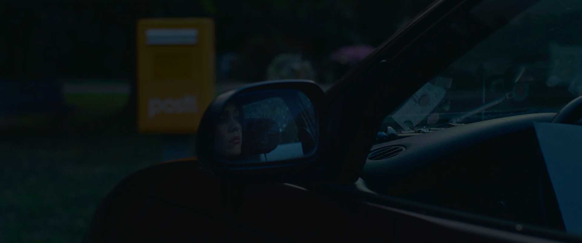 Close-up of a woman's reflection in the side mirror of a car, with a blurred yellow mailbox and forested background outside.
