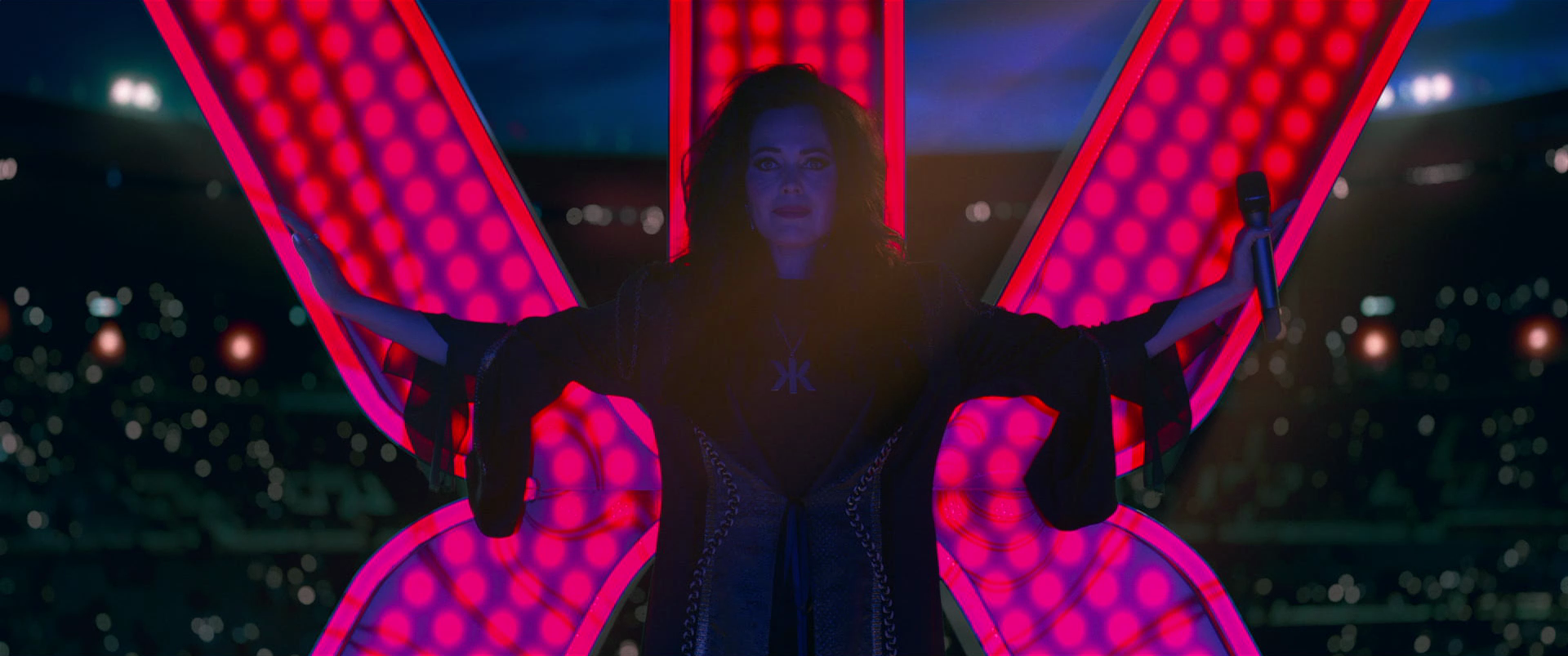 A woman with dark hair standing at night in front of illuminated pink and red butterfly-shaped lights, holding a microphone in her right hand, with city lights in the background.