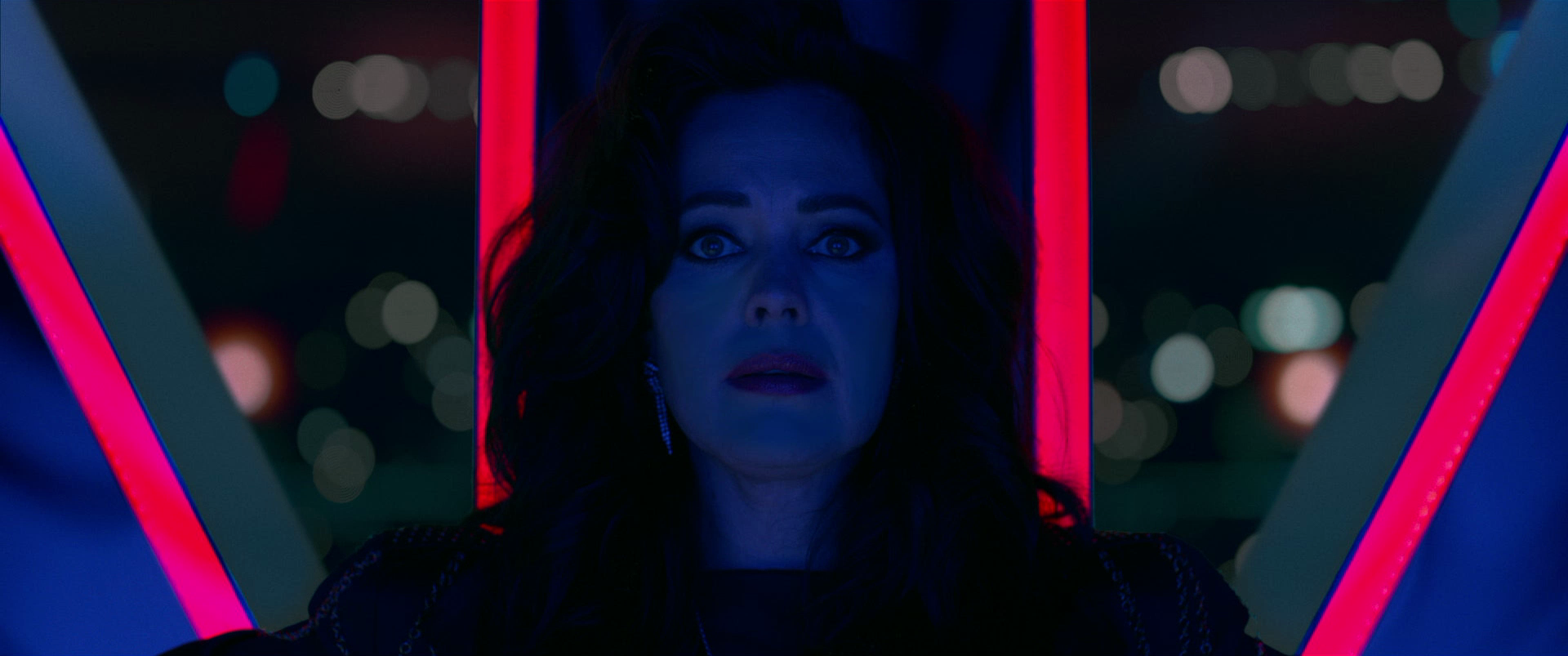 A woman with dark hair and earrings looks into a mirror with a surprised or shocked expression, illuminated by neon pink and blue lights, with a cityscape blurred in the background.