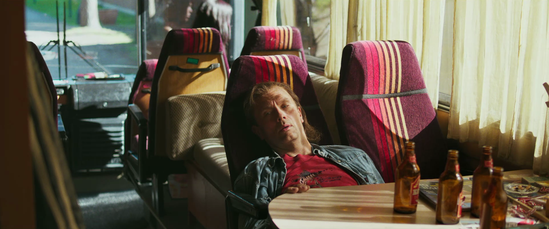 A man reclining on a bus seat with colorful, patterned fabric, looking at the camera. There are four bottles of beer on a table in front of him, and another person is visible in the background, sitting further back on the bus.