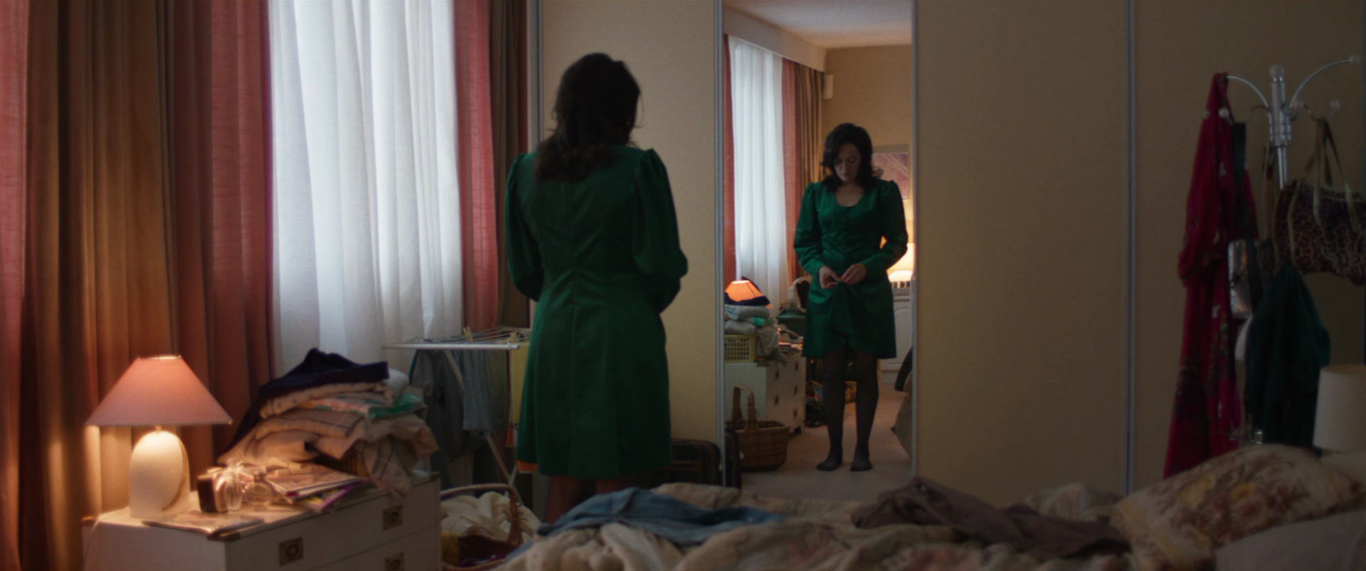 A woman in a green dress standing in front of a mirror, looking at her reflection in a bedroom. The room has a lit lamp, a pile of clothes, and a cluttered laundry area.