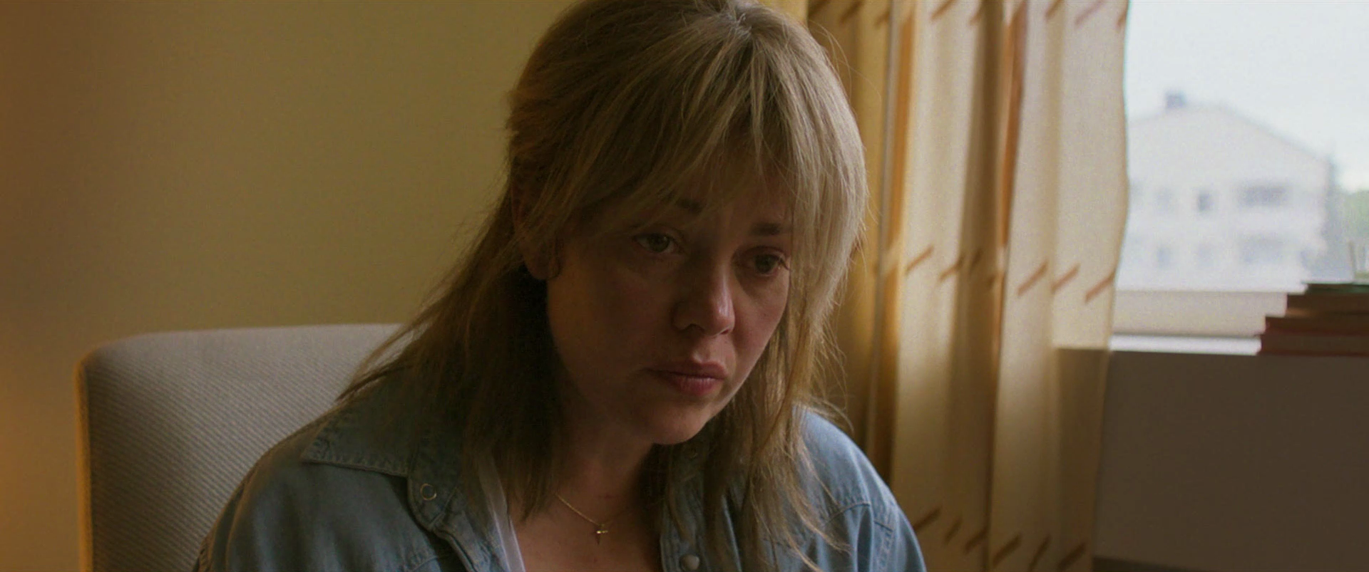 A woman with long, disheveled blonde hair sitting in a room with yellow curtains and a window, looking down with a serious or contemplative expression.