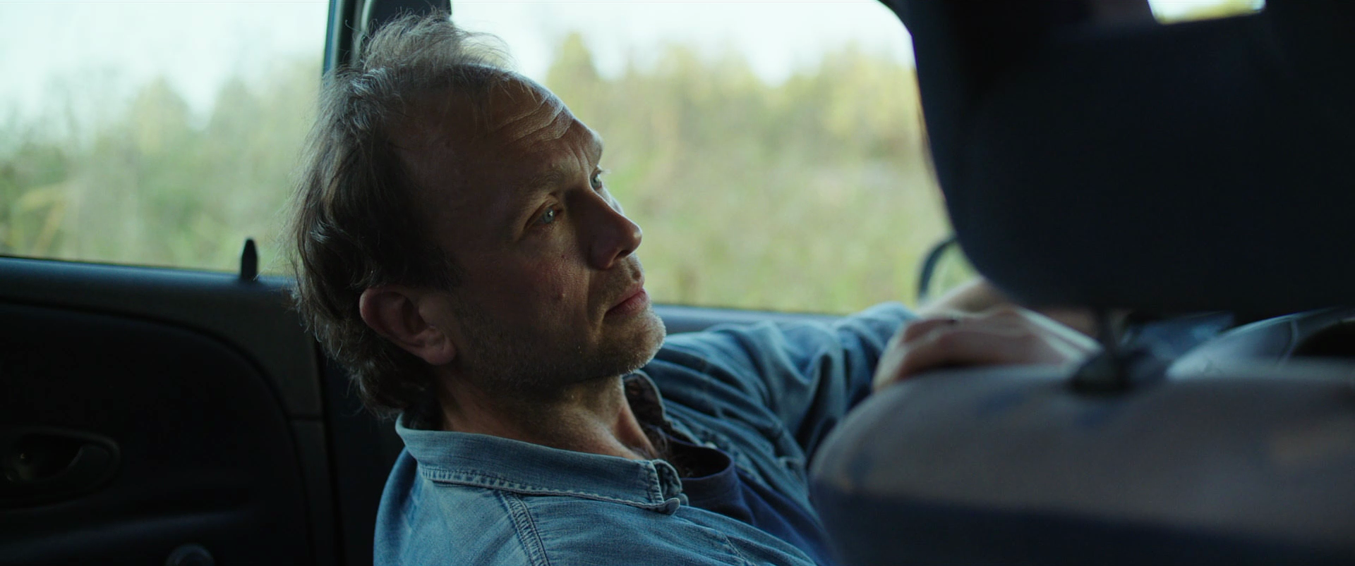 A man with light brown hair and a beard sitting in the backseat of a vehicle, looking thoughtfully out the window at a blurred, green outdoor landscape.