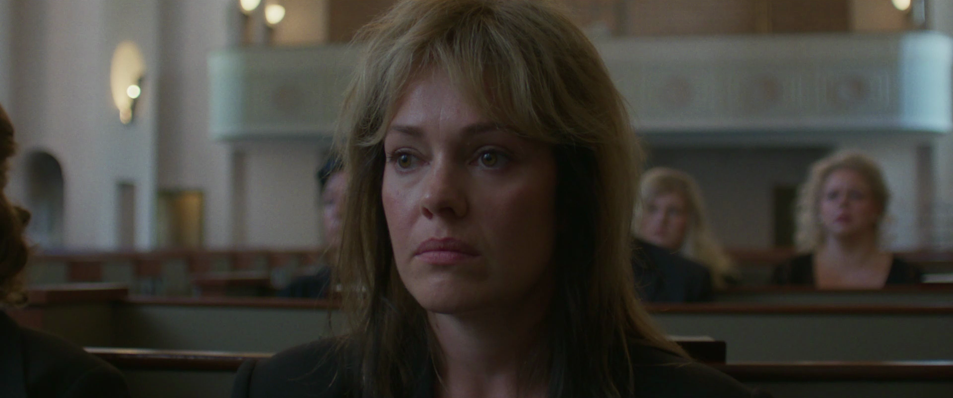 A woman with shoulder-length light brown hair and a serious expression sitting in a courtroom, with other women seated behind her.