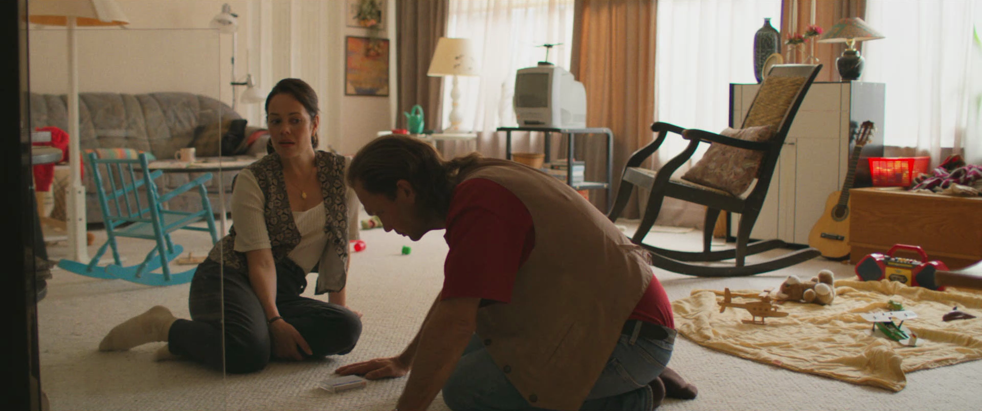 A woman and a man are inside a cluttered living room. The woman is sitting on the floor looking concerned, while the man is kneeling and reaching toward the carpet. Behind them, there are scattered toys, a rocking chair, a guitar, and other household