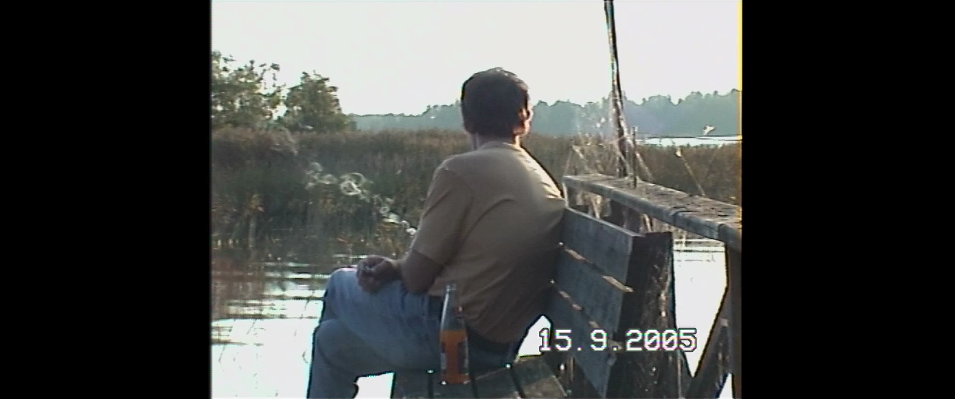 Man sitting on a park bench near water, smoking a cigarette, with a bottle of beer beside him, in a natural outdoor setting during late afternoon or evening.