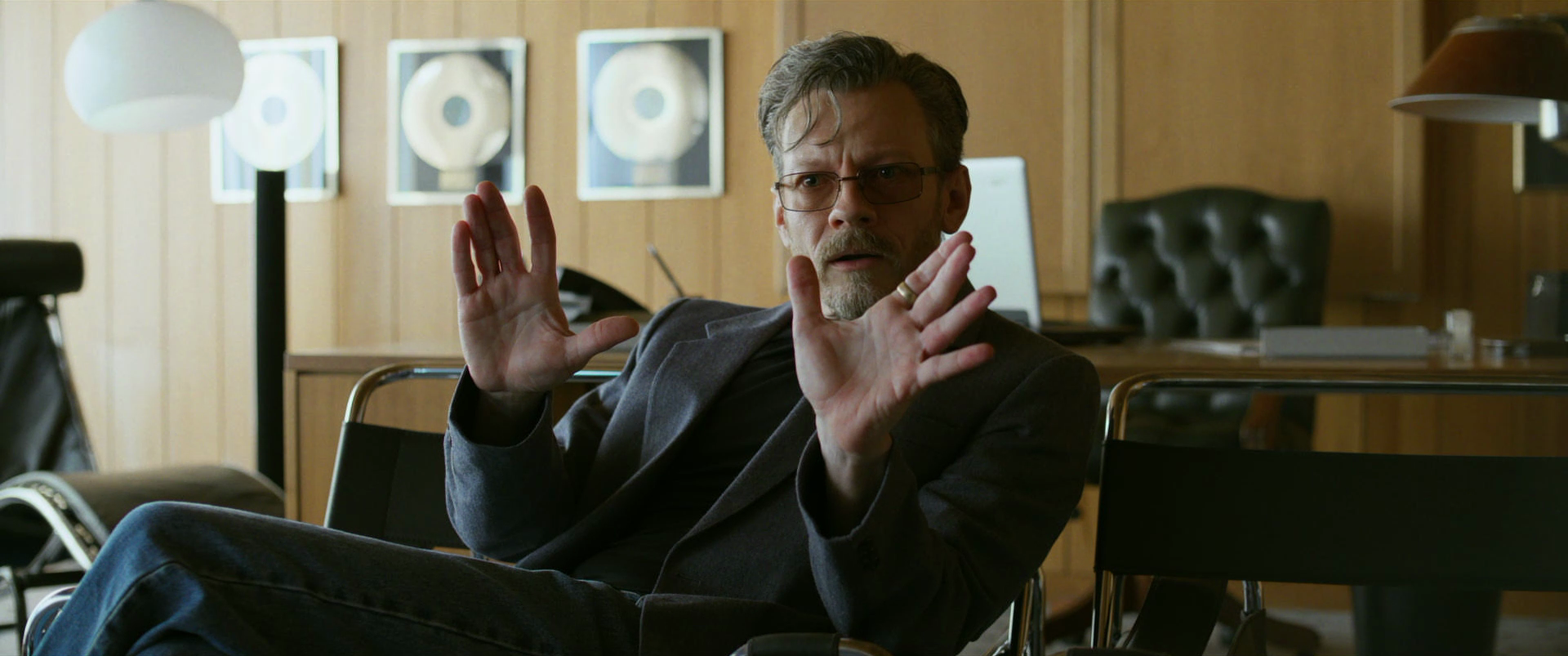 A man with glasses and a beard, wearing a dark blazer, is sitting on a chair and gesturing with his hands in an office setting, with framed gold records on the wall behind him.