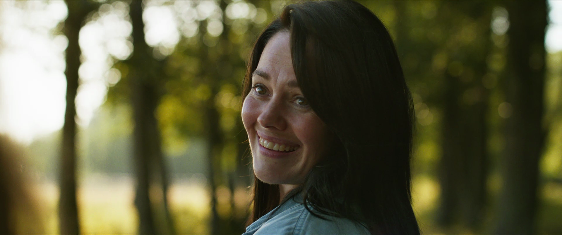 A woman with dark hair smiling outdoors in a forest setting with blurred trees in the background.