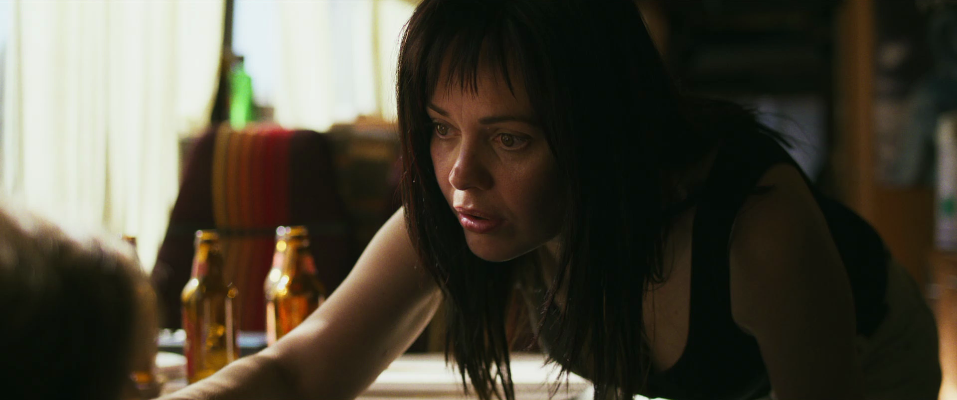 A woman with dark hair leaning forward, looking intensely at someone or something off-camera, with beer bottles on the table and a colorful striped cloth on a chair behind her, in a cozy room with soft lighting.