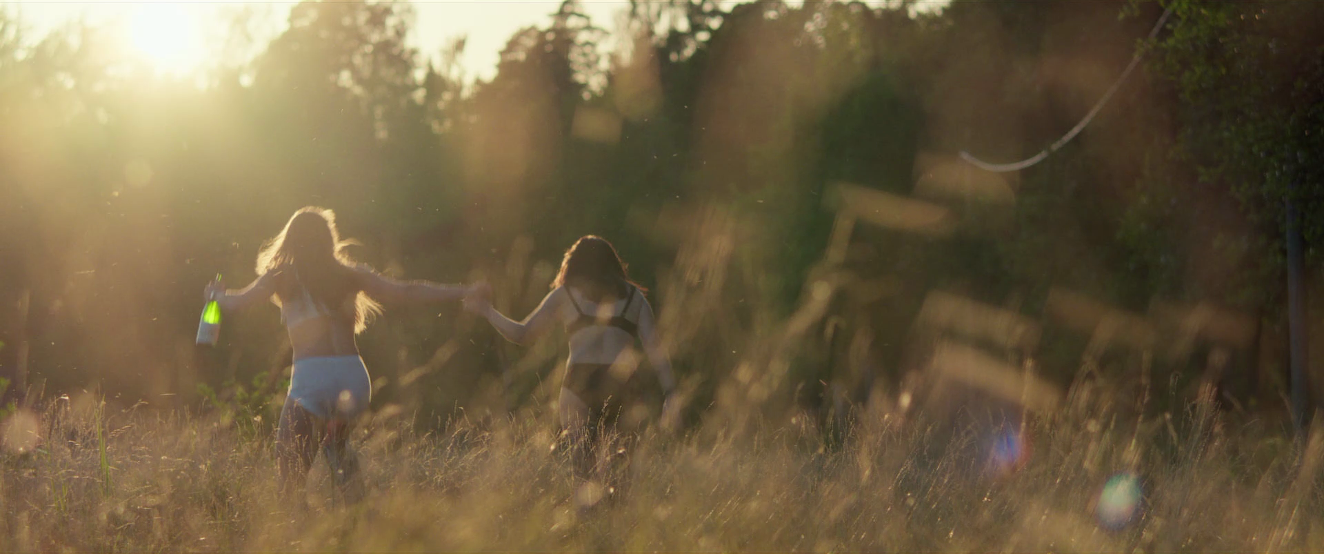 Two women in swimsuits holding hands and running through a grassy field at sunset, with trees in the background and sunlight creating a warm glow.