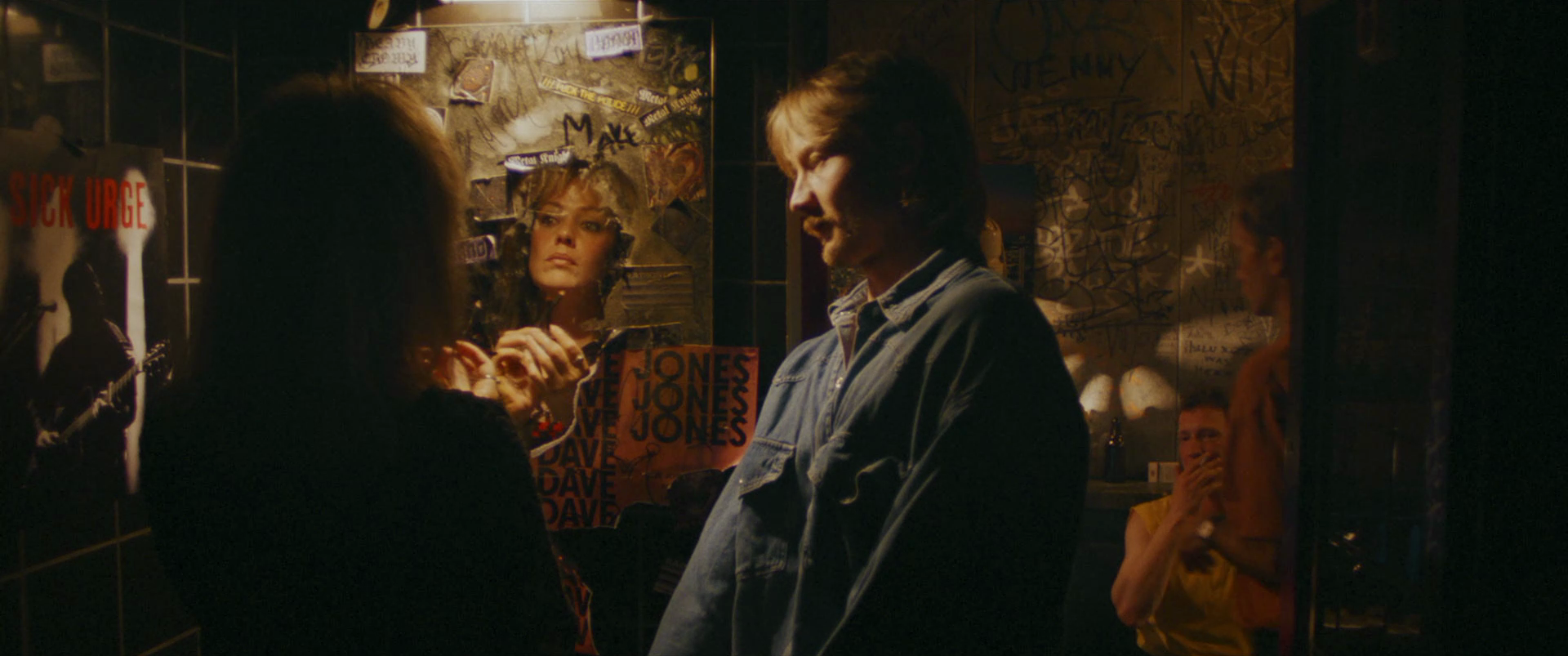 Scene in a dimly lit room with four people. In the foreground, a woman facing away from the camera and a man with long hair and a denim jacket. In the background, a woman looking in the mirror, and another woman sitting and smiling.