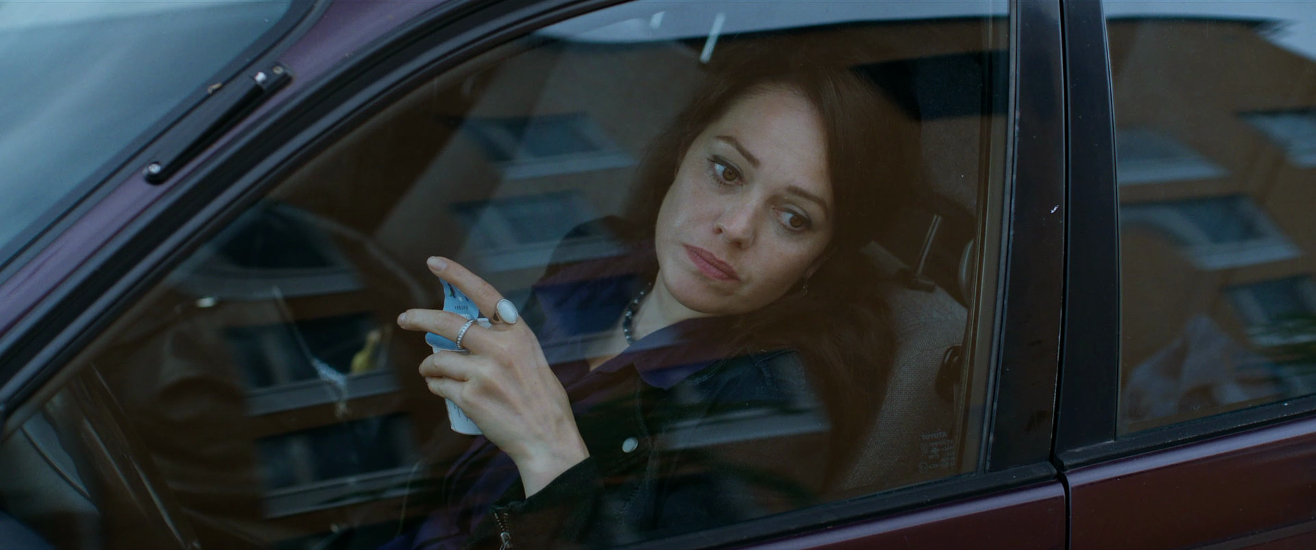 Woman with long dark hair sitting in the driver's seat of a car, viewed through the window, holding a note or ticket in her hand, with a contemplative expression.