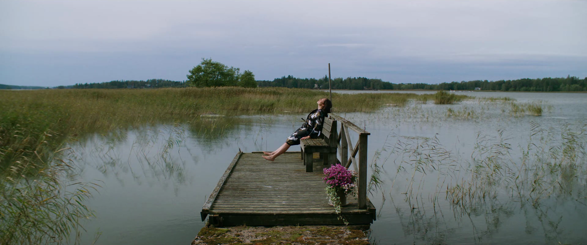 A woman sits on a wooden dock by a calm lake, leaning back with her eyes closed, enjoying the outdoors, surrounded by tall grasses and distant trees under an overcast sky.