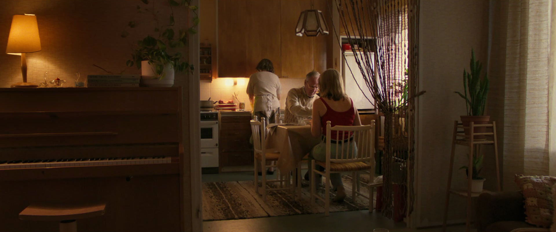 A cozy kitchen and dining area with four people. Two women sit at a table near a window, one woman stands at the stove, and a man is playing a game with a woman. Warm lighting, plants, and curtains add a homey atmosphere.