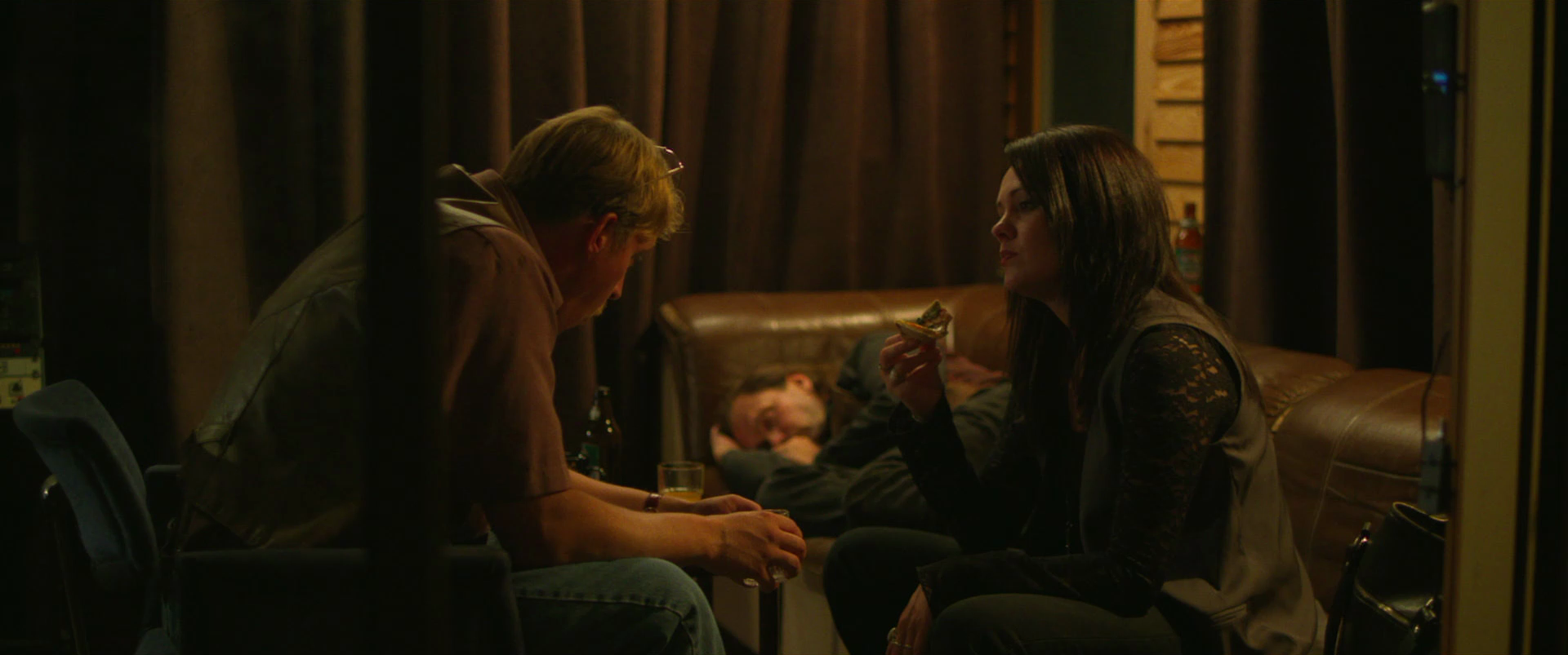 Two people sit and chat in a dimly lit room, one holding a slice of pizza, with a third person resting on a couch in the background.