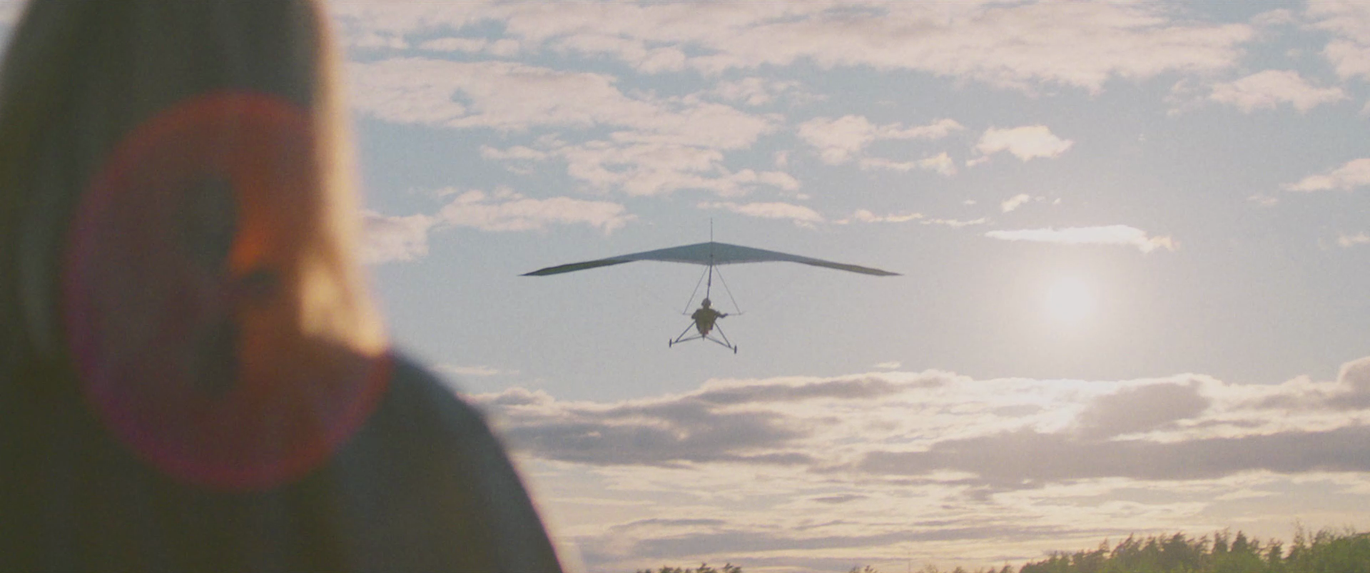 A helicopter flying in the sky with clouds and the sun shining, viewed from the ground where a blurred figure is partially visible on the left.