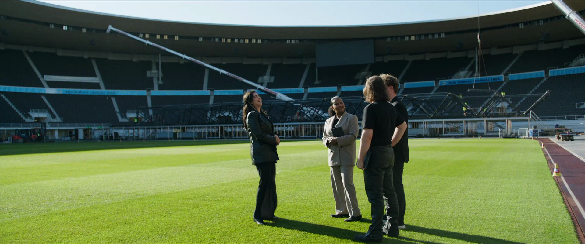 Group of four people standing and talking on a football field inside a stadium during daylight. They are wearing formal and business casual attire, and the stadium appears to be under construction or maintenance.