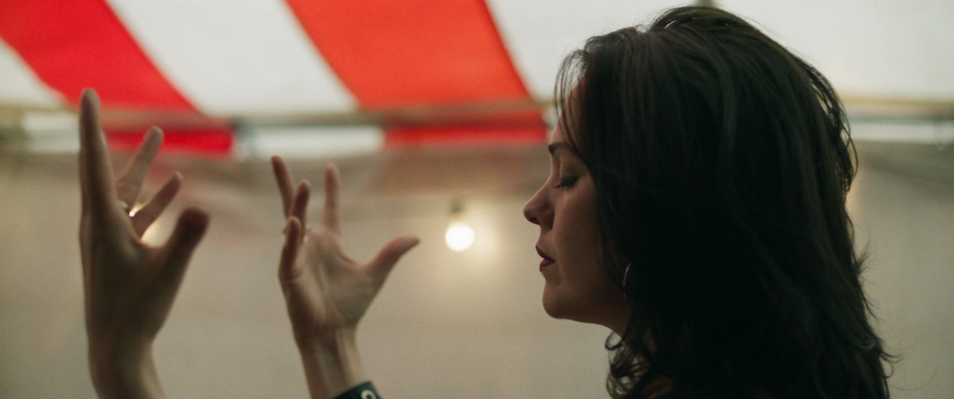 A woman with dark hair and earrings is seen in profile, with her eyes closed, as she raises her hands with fingers spread near what appears to be a red and white striped tent or decoration.