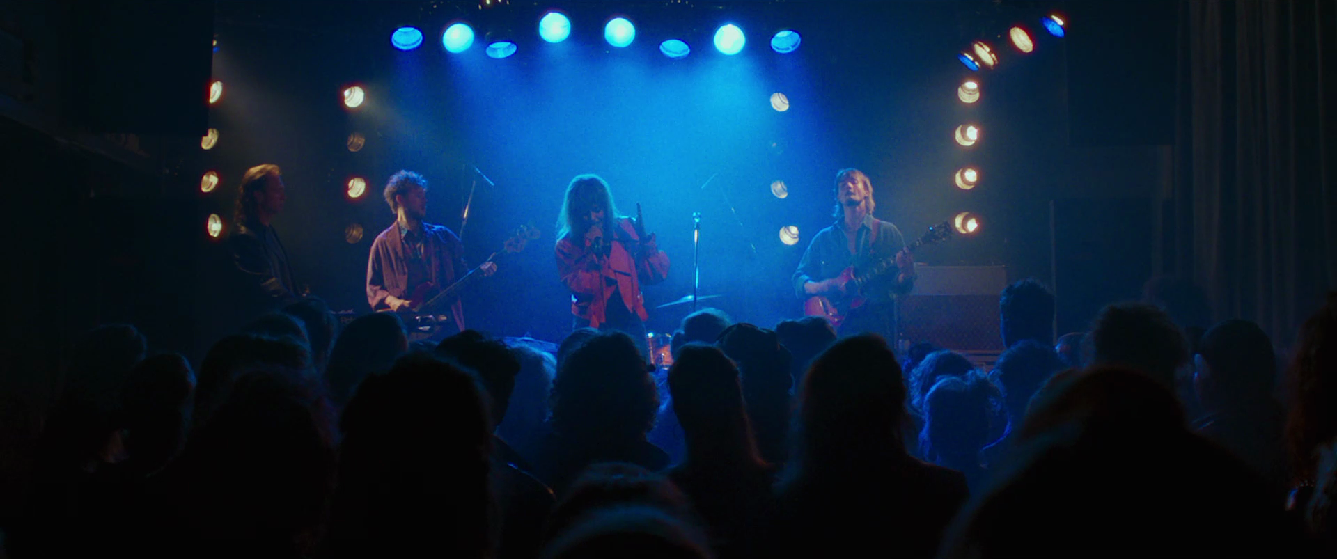 A live music performance on stage with four band members playing instruments and singing, illuminated by colorful stage lights, with an audience watching.