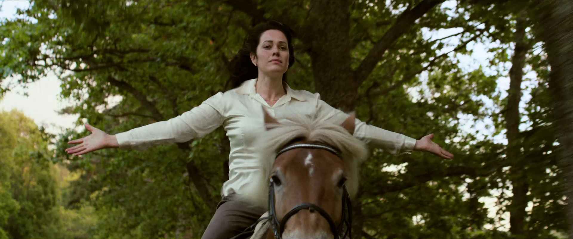 A woman with dark hair, wearing a light-colored blouse, is riding a horse with arms outstretched in a green, leafy outdoor setting.