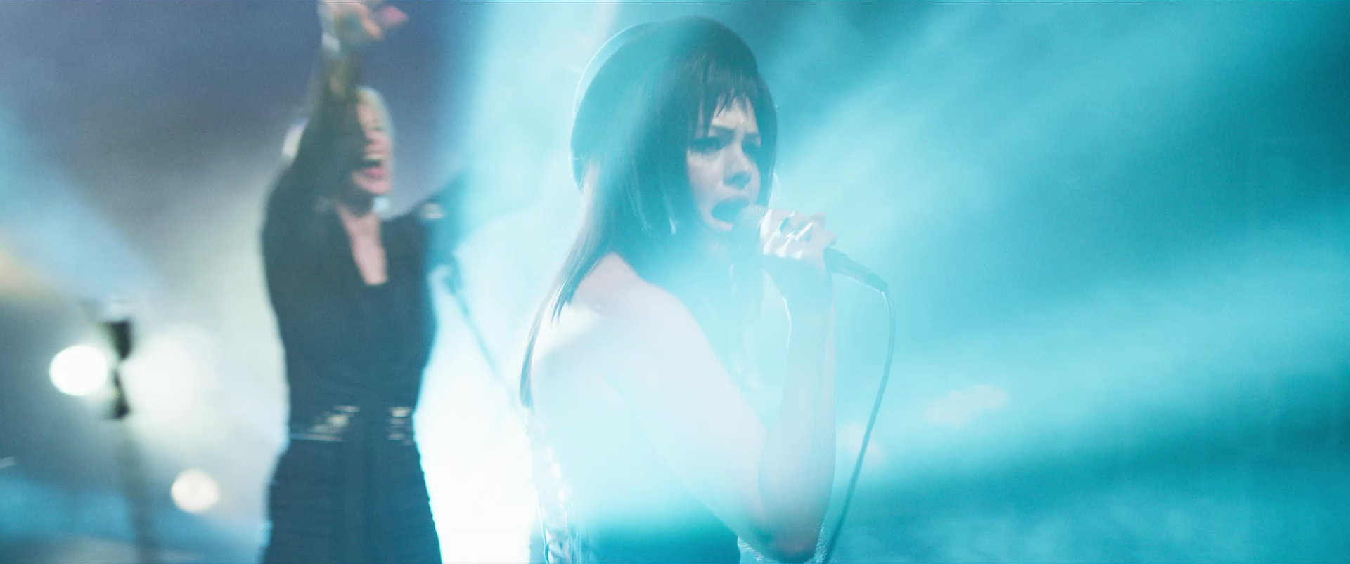 Two women singing into a microphone on stage, surrounded by blue stage lighting and smoke effects.
