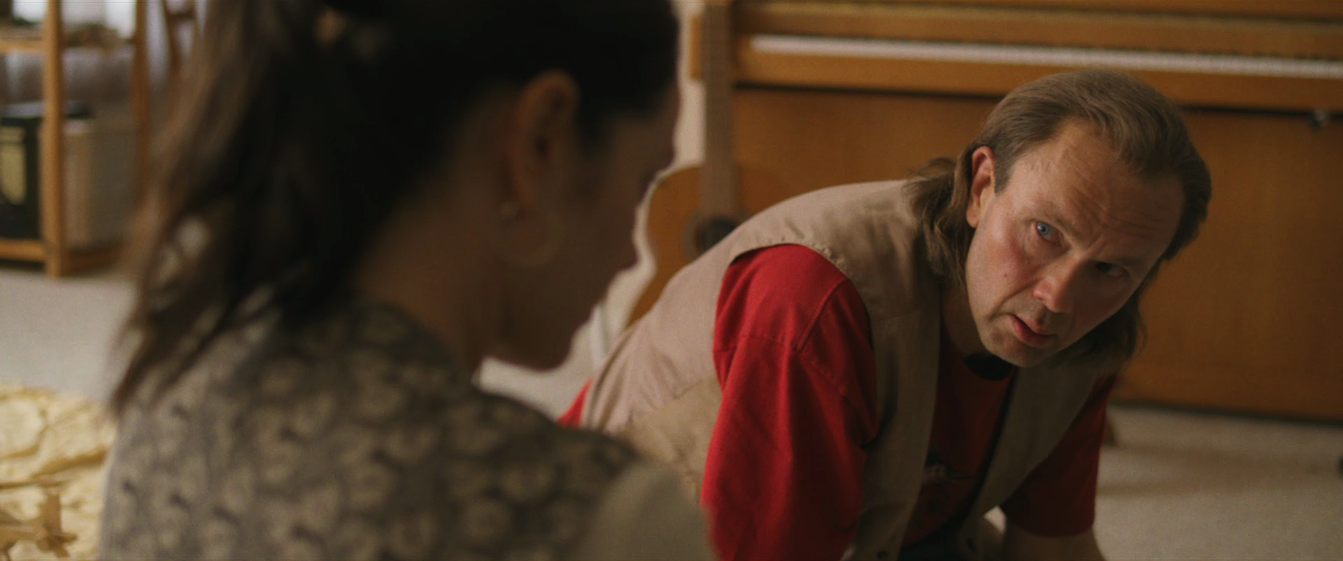 A man with long hair and a red shirt, possibly with a beige vest, is looking at a woman with dark hair and hoop earrings. They are sitting close together indoors, with wooden furniture in the background.