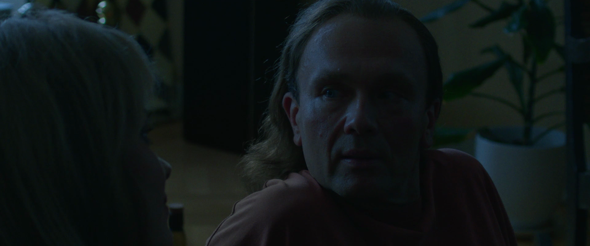 A dark scene with two people, a man with shoulder-length hair and a woman with blonde hair, engaged in a conversation. The man is looking at the woman with a serious expression, and there are plants in the background.