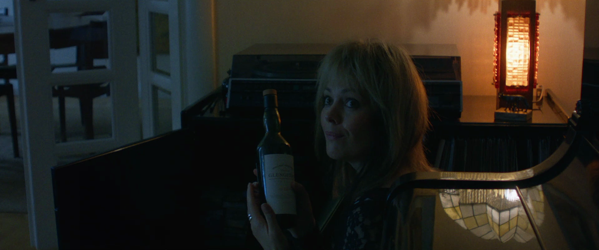 A woman with long blonde hair holding a wine bottle and looking towards the camera, sitting in a dimly lit room with a decorative lamp on a table in the background.