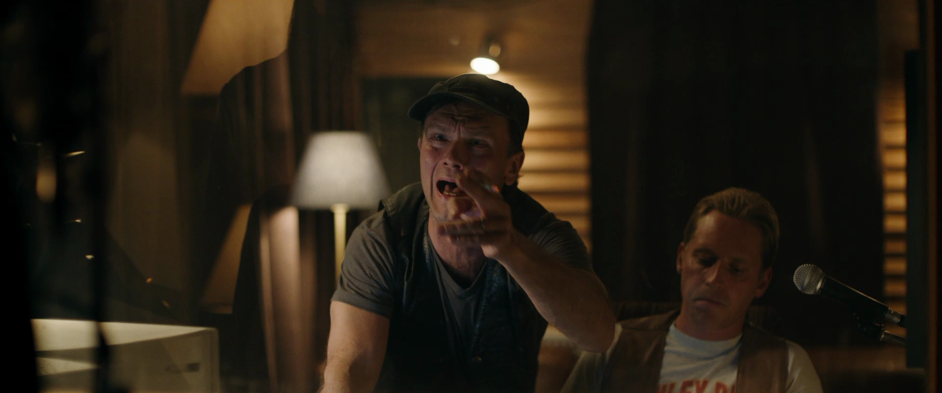 Two men are in a dimly lit room with wooden walls and furniture. One man is standing, shouting, and gestures with his hand, while the other man is sitting and appears to be focused, with a microphone nearby.