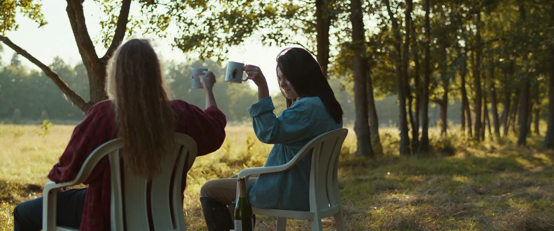Two women sitting outdoors in a wooded area, raising coffee mugs in a toast during sunset, with a bottle of champagne on the ground.
