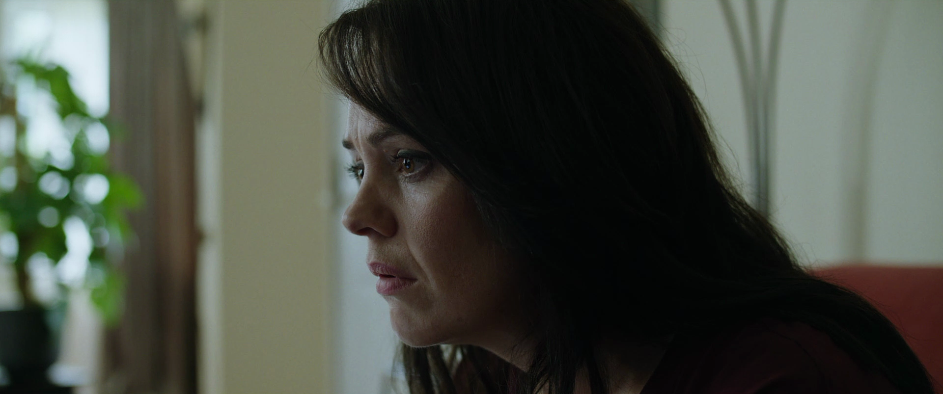 A woman with dark hair looking contemplative, sitting indoors with a blurred green plant and window in the background.
