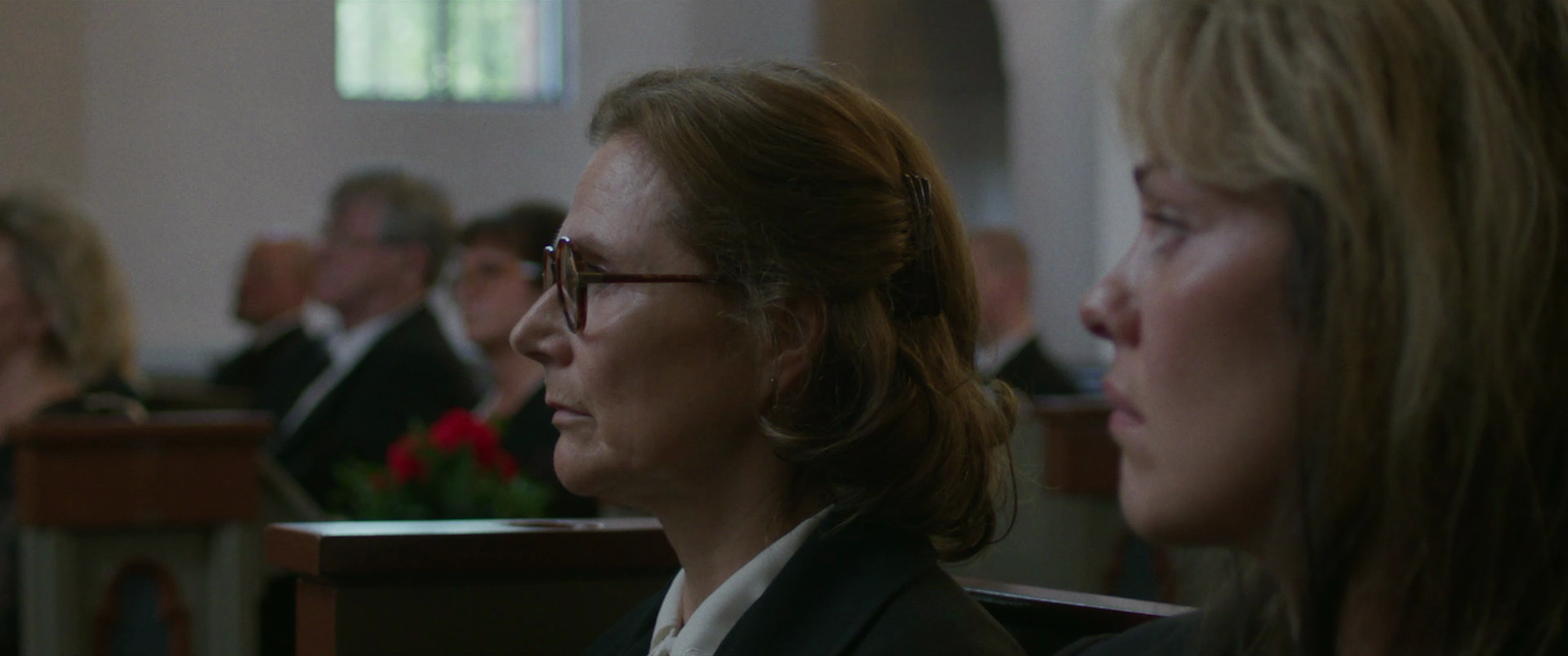 Side profile of two women sitting in a church or funeral setting with somber expressions, with other attendees visible in the background.