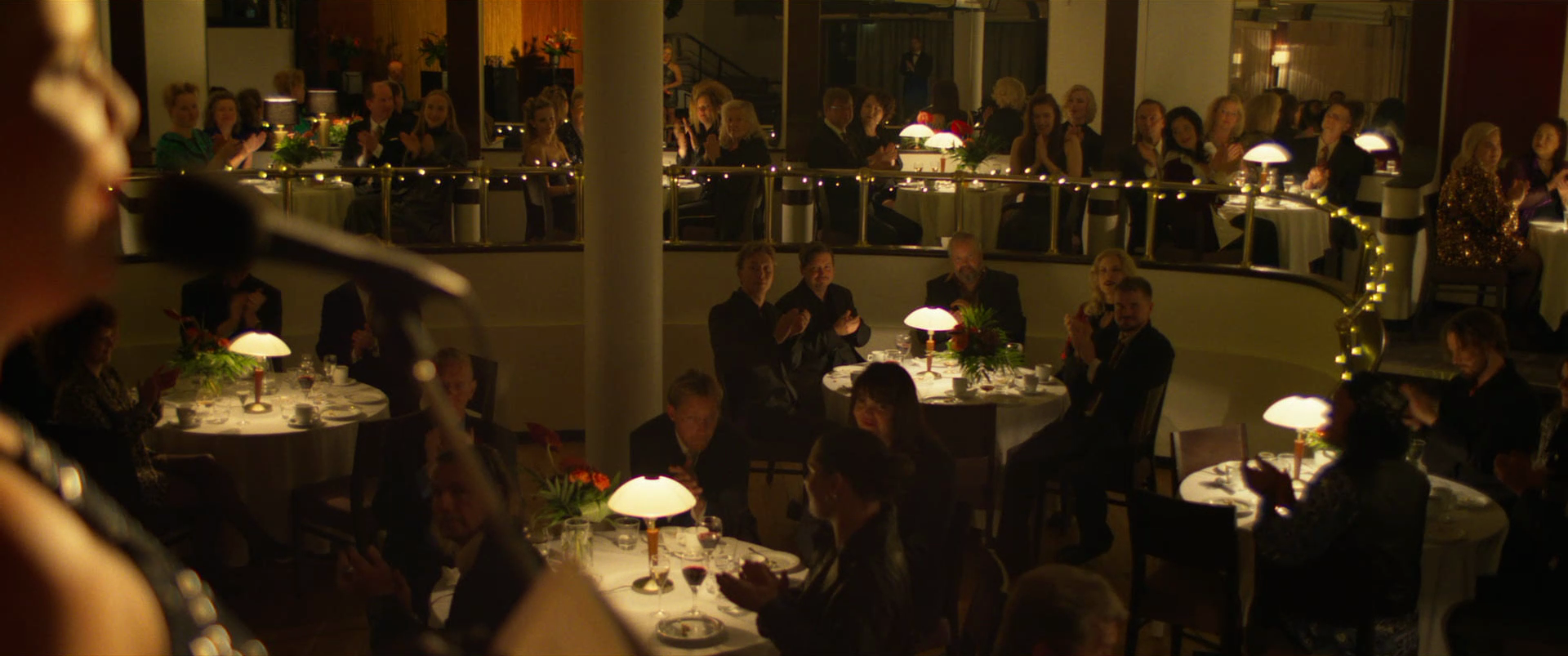 People sitting at round tables with white tablecloths, flowers, and lamps in a dimly lit banquet hall or restaurant, clapping and watching a speaker or performer.