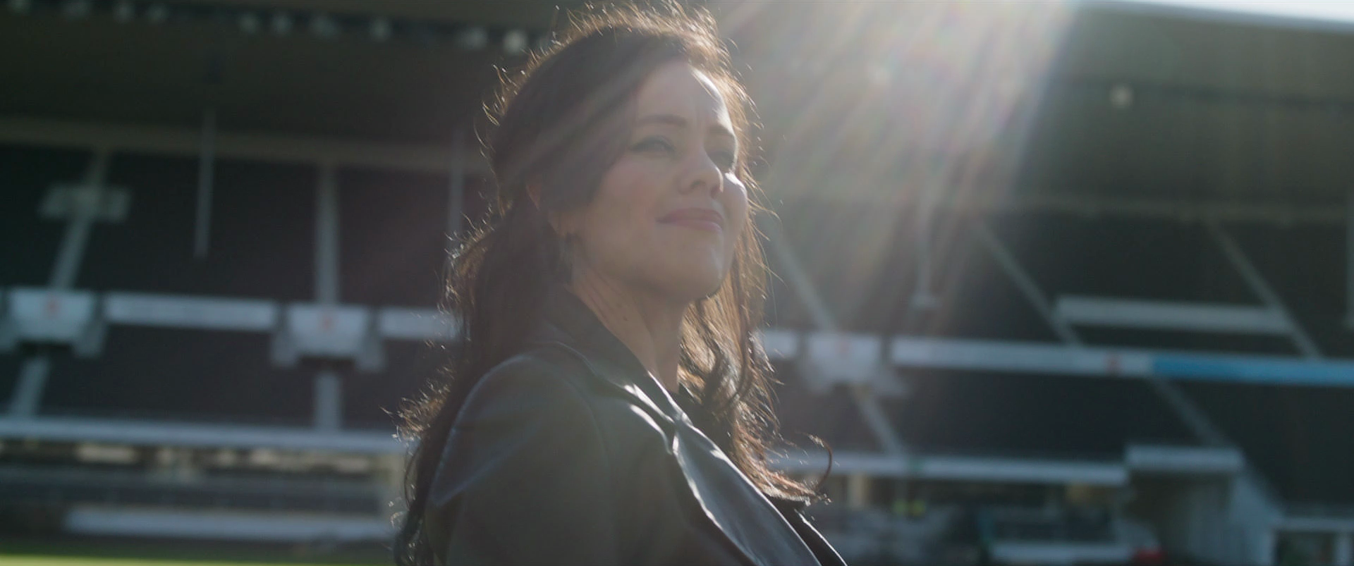Smiling woman with dark hair, wearing a leather jacket, standing outdoors in front of stadium bleachers with sunlight.