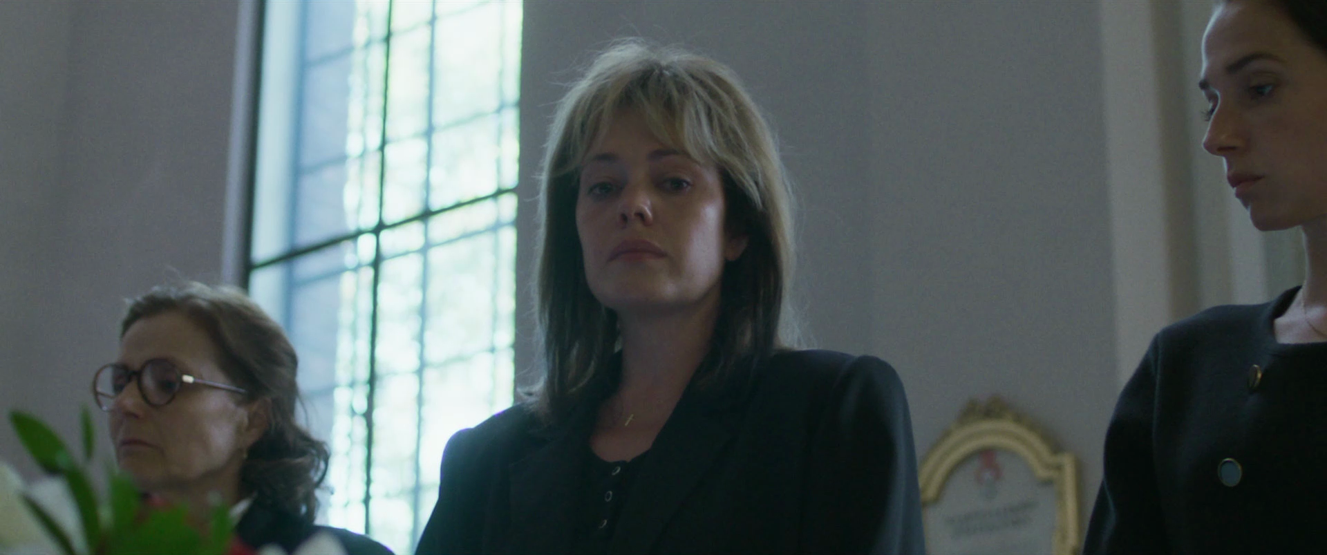 Three women sitting at a table in a room with large window. The woman in the center has shoulder-length blond hair and a serious expression. The woman on the left has glasses and darker hair, looking down. The woman on the right has dark hair pulled 