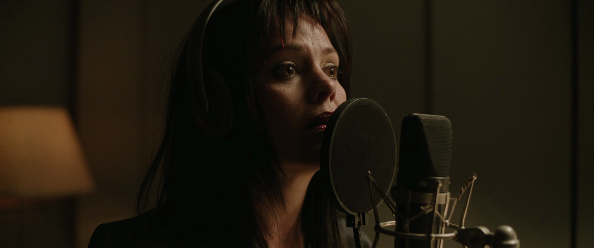 A woman is singing into a microphone in a recording studio, wearing headphones with a dimly lit background.