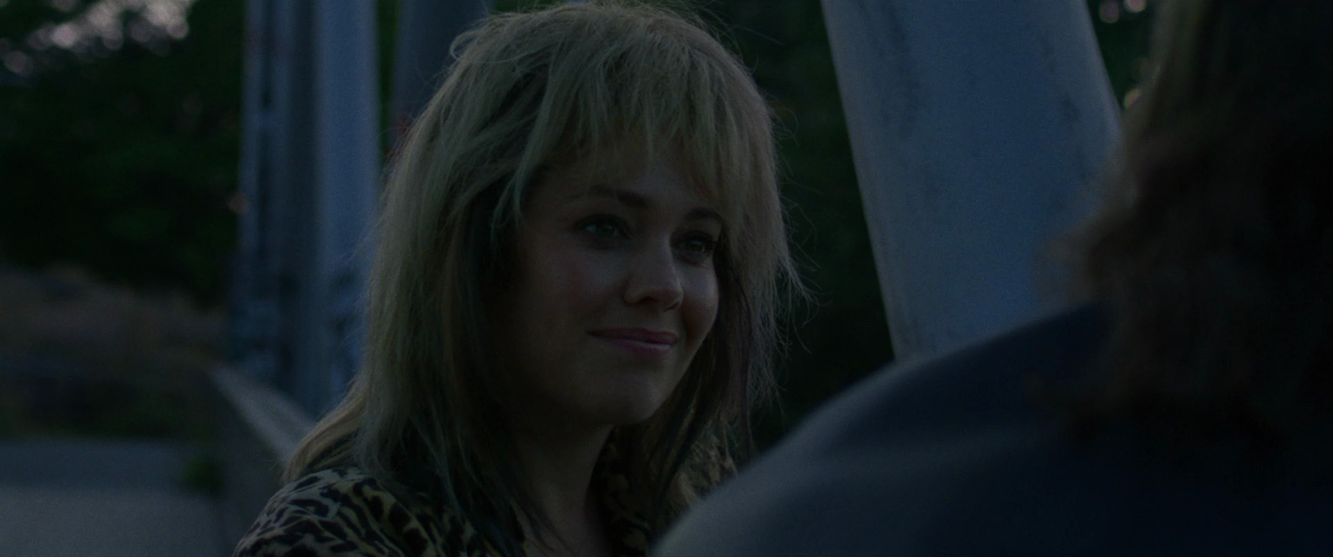 A woman with blonde, messy hair smiling while talking to someone outside during the evening.